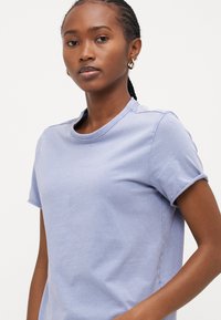 Tricou din bumbac de culoare mov deschis, cu mâneci scurte, decolteu rotund și manșete răsucite. Are o textură simplă și netedă și fiecăre parte cu șanțuri laterale.