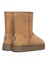 Botas de tobillo de ante marrón con forro suave, suelas de goma marrones resistentes y una etiqueta con el logo en la parte trasera. Costuras visibles.