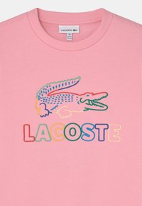 Sweat-shirt rose avec un logo crocodile brodé au-dessus du mot "LACOSTE" en lettres multicolores. Texture lisse et col rond.