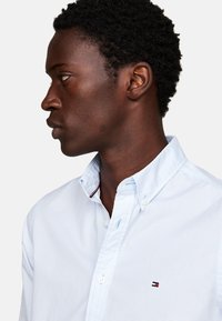 Camisa de botones azul claro con un pequeño patrón de cuadros, botones blancos y un detalle de logo en el pecho. Con cuello y un acabado pulcro.