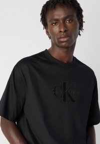 Crna pamučna majica s kratkim rukavima, s reljefnim logotipom "Calvin Klein" na prednjoj strani. Glatka tekstura i standardni dizajn s okruglim izrezom.