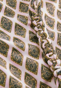 Tekstil med et gentaget diamantmønster i guld og blå på en lyserød baggrund, med en flettet accent i matchende farver.