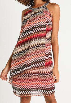 Mujer con vestido sin mangas hasta la rodilla con patrón de zigzag colorido en rojo, rosa, marrón, beige y negro, con escote adornado.