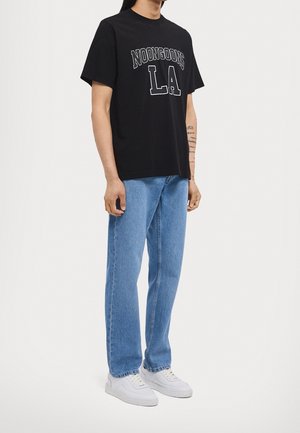 Person iført en sort T-shirt med teksten "NOONGOONS LA", lyseblå jeans, hvide sneakers, stående mod en ensfarvet lys baggrund.