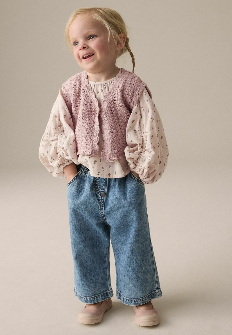 Lichtroze gebreide vest over een langlopende blouse met bloemenpatroon, gecombineerd met losse blauwe denim jeans en lichtgekleurde schoenen.