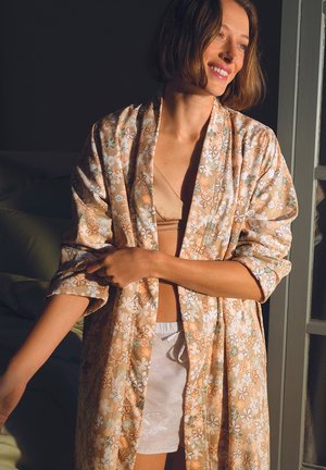 Femme aux cheveux bruns courts souriante, portant une robe fleurie sur un soutien-gorge beige et un short blanc, debout près d'une fenêtre baignée de lumière solaire.