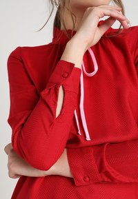 Czerwona bluzka z teksturowanym wzorem w jodełkę, z wysoką szyją, różowym paskiem ściągającym i mankietami zapinanymi na guziki. Gładki materiał, dopasowany krój.