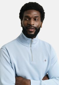 Hellblaue Halb-Zip-Sweatshirt aus weichem Material, mit dekorativem Reißverschluss und dezentem Markenlogo auf der Brust. Strukturierte Oberfläche.