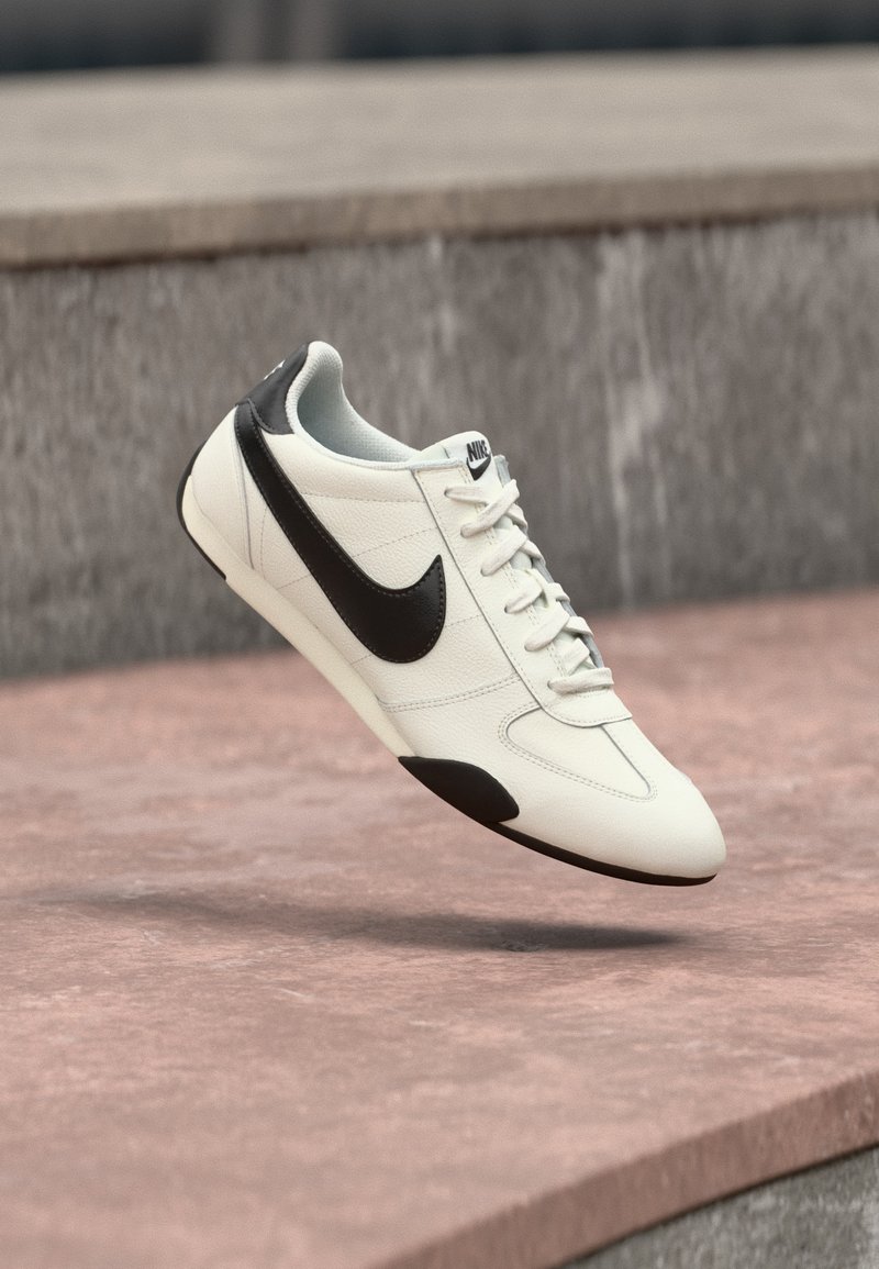 Weißer Nike-Sneaker mit schwarzem Swoosh-Logo, schwarzen Akzenten an Ferse und Sohle, schwebt über einer Steinfäche im Freien.