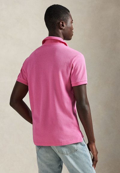 Πόλο με κοντά μανίκια από βαμβακερό μείγμα σε ζωηρό ροζ χρώμα, με κλασικό γιακά και πλαϊνές σχισμές, φορεμένο με ανοιχτό μπλε τζιν παντελόνι.