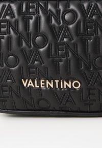 Černá kožená taška s vyraženým vzorem názvu "VALENTINO" a zlatým kovovým logem. Hladký povrch, obdélníkový tvar.