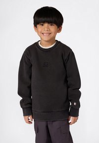 Ragazzo sorridente con capelli neri, indossa un felpa nera Champion e pantaloni cargo scuri, in piedi contro uno sfondo bianco.
