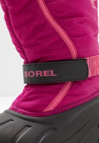 Nära bild av en rosa vinterstövel med svart sula och justerbart svart rem med etikett "SOREL" på sidan.