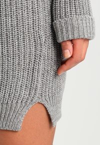 Handen vilar mot låret och bär en grå ribbad stickad tröja med uppvikta ärmar och en liten spricka på sidan vid fållen.