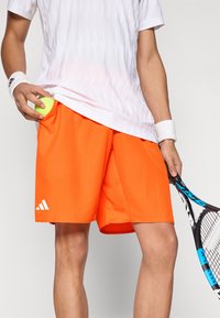 Helderoranje sportshorts met een mesh-ontwerp, met het witte Adidas-logo en zijzakken. Het model houdt een gele tennisbal vast.