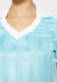 Ljusblå Adidas sporttröja med vit krage och logo på bröstet, bärs av en person som visas från halsen till mitten av överkroppen.