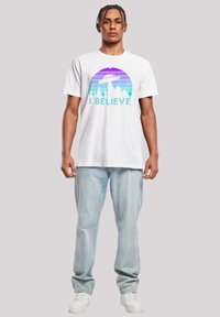 White cotton t-shirt featuring a colorful graphic of mountains and a UFO, with the text "I BELIEVE" in turquoise. Paired with light blue jeans.