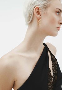 Pendiente en forma de espiral dorada; diseño suave y ligero; usado por un modelo con el cabello corto; combinado con una prenda negra con detalles de encaje.