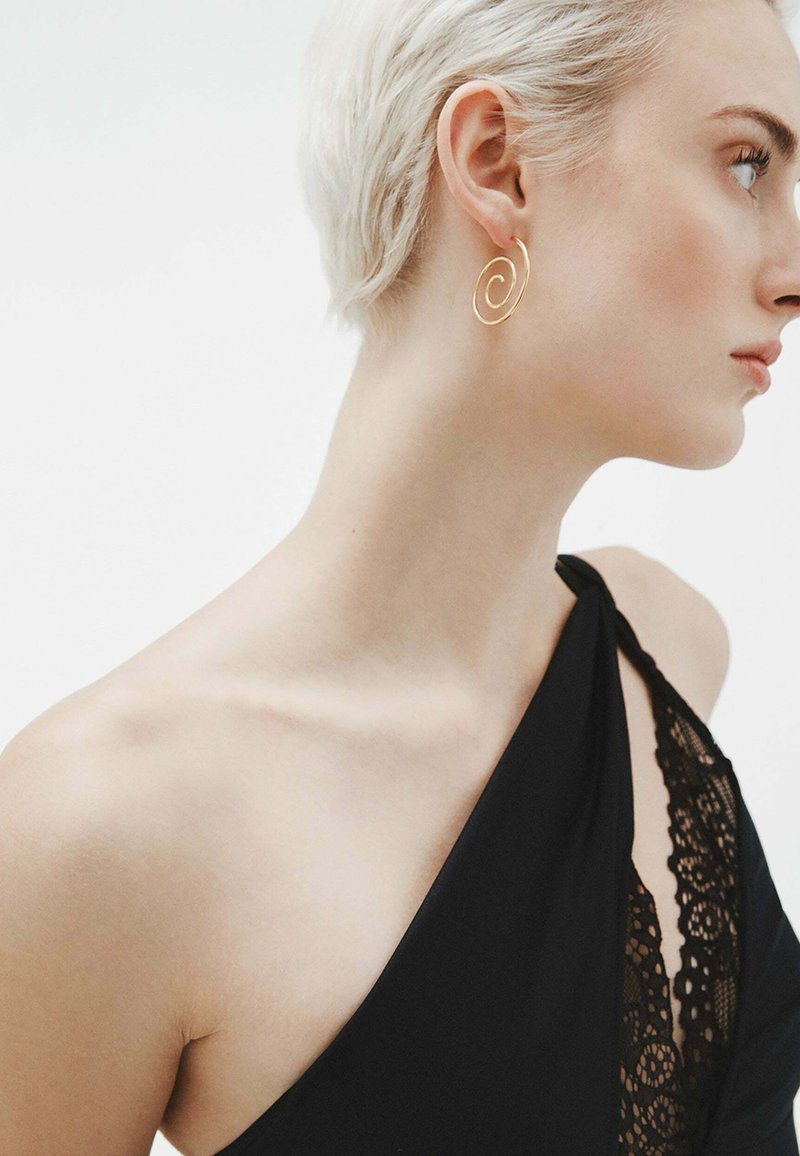 Pendiente en forma de espiral dorada; diseño suave y ligero; usado por un modelo con el cabello corto; combinado con una prenda negra con detalles de encaje.