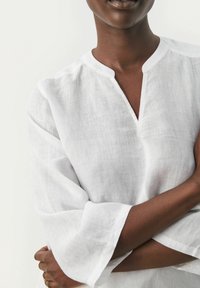 Personne avec la peau foncée portant un haut en lin blanc avec un décolleté fendu, les bras croisés sur le torse contre un fond clair.