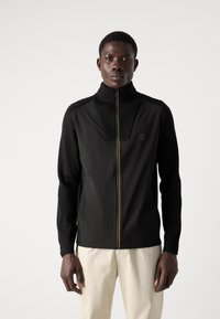 Jachetă neagră cu fermoar, combinând un material neted cu un tricot texturat. Are un guler înalt, fermoar auriu și un detaliu cu logo pe piept.