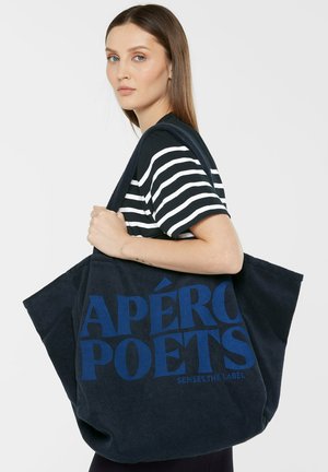 Junge Frau in schwarz-weiß gestreiftem Shirt hält große dunkelblaue Tragetasche mit dem Text "APÉRO POETS SENSES.THE LABEL".
