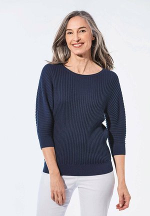 Marineblauer gerippter Strickpullover mit weitem Rundhalsausschnitt, dreiviertellangen Ärmeln und strukturierten Details entlang der Ärmel. Kombiniert mit weißen Hosen.