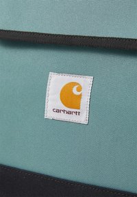 Sac à dos en tissu teal avec des accents noirs, comportant une étiquette en tissu blanc affichant un logo orange. Le matériau a une finition texturée.