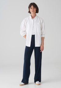 Witte overhemd met lange mouwen, gedragen over een wit T-shirt. Gecombineerd met marineblauwe wijde broek en witte sneakers.