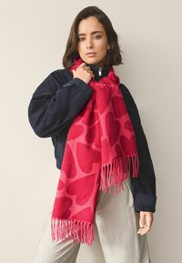 Bufanda de punto con base roja y un patrón de corazones en diferentes tonos de rojo, terminada con flecos en los extremos.