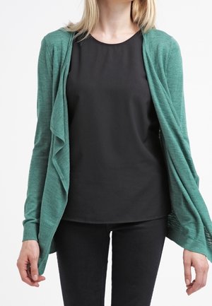 Tefkleurige cardigan met een open voorkant, lichte textuur en gedrapeerd ontwerp. Gemaakt boven een zwarte top en gecombineerd met zwarte broeken.