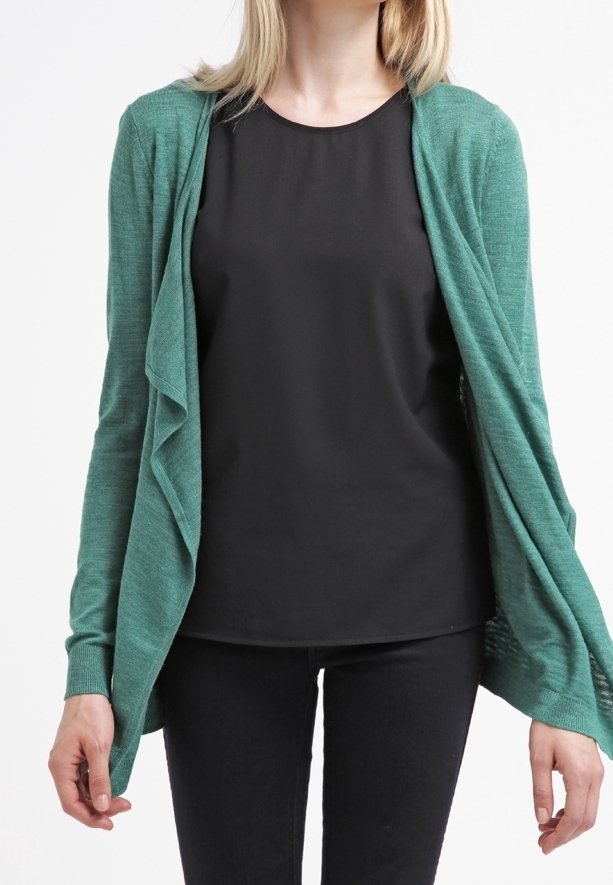 Vihreä karjakauluksellinen neuletakki, jossa on avoin etuosa, kevyt rakenne ja roikkuva muotoilu. Puettuna mustan paidan päällä ja yhdistettynä mustiin housuihin.