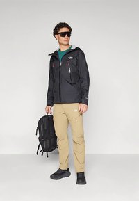 Hombre con chaqueta negra con capucha, pantalones beige y zapatos negros, sosteniendo una mochila negra y llevando gafas de sol grandes y oscuras.