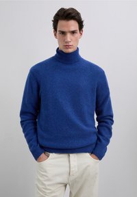 Maglione a collo alto blu lavorato a maglia con una finitura testurizzata. Il maglione presenta polsini e orlo a coste, abbinato a pantaloni beige chiaro.