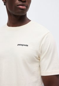 Κοντινό πλάνο ενός ατόμου που φοράει ένα εκρού t-shirt Patagonia με το λογότυπο τυπωμένο μαύρο στο αριστερό μέρος του στήθους.