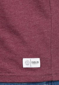 Burgundy szövet, rajta egy fehér, téglalap alakú ruhacímke, amelyen az "!SOLID DENMARK" felirat olvasható, a nadrág alsó szegélye felett, kék farmer mellett.