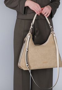 Beżowa torebka z zamszu z kremowymi akcentami, o strukturalnym kształcie i jednej rączce. Posiada zamek błyskawiczny i złote detale z logo.