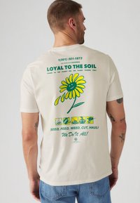 T-shirt en coton crème avec un graphique de fleur jaune arborant un visage souriant, du texte vert, et plusieurs petites icônes en bas.