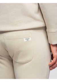 Pantalon de survêtement beige avec poche arrière et petite étiquette blanche portant l'inscription « YOUR ONLY LIMIT IS YOU AND », porté par une personne aux doigts tatoués.