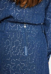 Denim tkanina s srebrnimi bleščicami, z teksturiranim vzorcem. Na sprednji strani je vidna zadrga z modrim zategovalnikom.
