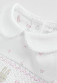 Hvid beklædningsgenstand med en bølget krave, dekoreret med broderede kanin- og blomster-motiver. Blødt stof med struktureret smock og lyserøde detaljer.