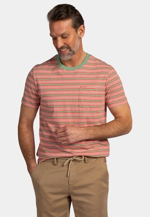 Man draagt beige broek en een gestreept T-shirt met korte mouwen met groene en rode horizontale strepen, kijkt naar beneden en glimlacht.