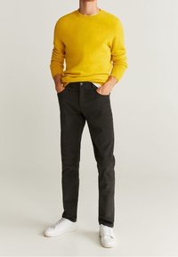 Gelber gestrickter Pullover mit gerippten Bündchen, kombiniert mit schwarzen Slim-Fit-Hosen und weißen Turnschuhen. Schlichtes Design, keine sichtbaren Muster oder Akzente.