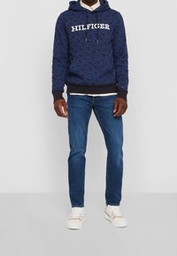 Tmavě modrá mikina s vzorem a logem "HILFIGER", zkombinována s světle modrými džíny a bílými teniskami s béžovými akcenty.
