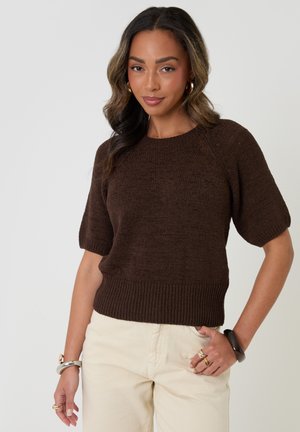 Frau trägt einen dunkelbraunen kurzärmeligen Strickpullover und hellbeige Hose, mit goldenen Creolen und Armbändern an beiden Handgelenken.