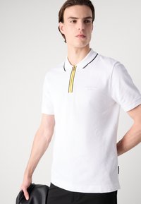 Baltais īsām piedurknēm polo krekls, izgatavots no kokvilnas, ar dzeltenu rāvējslēdzēju un melniem akcentiem, kā arī smalku logotipu uz krūtīm.