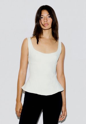 Femme aux longs cheveux foncés portant un haut blanc ajusté sans manches et un pantalon noir, debout devant un fond clair uni.