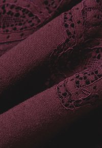 Burgundowa tkanina o miękkiej strukturze, z misternymi detalami koronkowymi i jednolitym, tkanym wzorem.