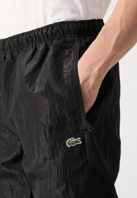Schwarze Nylonschorts mit elastischem Bund, seitlichen Taschen und einer strukturierten Oberfläche. Mit einem kleinen grünen Krokodil-Logo als Akzent.