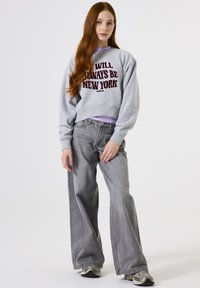 Sudadera gris corta con texto burdeos "SIEMPRE SERÉ NUEVA YORK" sobre una camiseta de manga larga lavanda, combinada con jeans grises de pierna ancha.